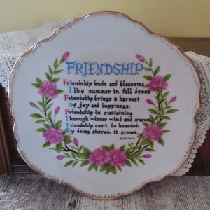 May include: White decorative plate with a scalloped edge and gold trim. The plate features the word "FRIENDSHIP" in blue script, surrounded by a floral wreath of pink roses and green leaves. A poem about friendship is written on the plate.