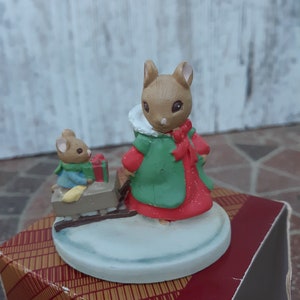 May include: A Christmas figurine set featuring two brown mice. The larger mouse is wearing a green coat with a red trim and a red bow. The smaller mouse is pulling a red sled with a wrapped gift. The figurine is sitting on a white base.