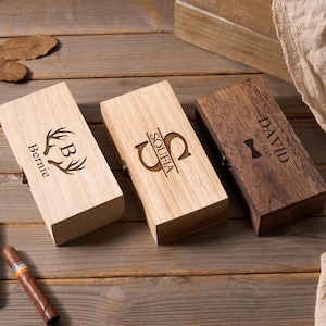 以下が含まれることがあります： パーソナライズされた彫刻が施された3つの木製ボックス。1つのボックスは明るい色で、「Bernie」と鹿の角のデザインが施されています。もう1つは「Soufia」と刻まれています。3つ目の、より暗いボックスには「David Best Man」と書かれています。2本の葉巻が見えます。