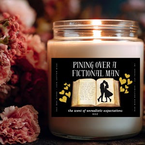 May include: A lit candle in a glass jar with a black label. The label reads "Pining Over A Fictional Man" with an illustration of a couple kissing over an open book. Gold heart accents surround the book. The candle is surrounded by pink and red flowers.
