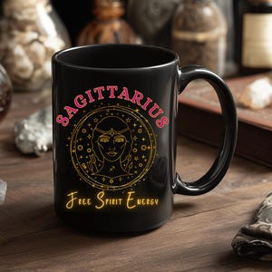 Peut inclure: Mug en céramique noire avec le mot "SAGITTARIUS" en rose, entourant une illustration dorée d'un visage de femme. En dessous, les mots "Free Spirit Energy" sont écrits en jaune. Le mug est posé sur une surface en bois.