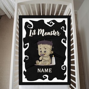 May include: Black baby blanket with a white border featuring a cartoon Frankenstein's monster baby design. The text "Lil Monster" is at the top, with space for a name at the bottom. A pacifier and bottle are also included.