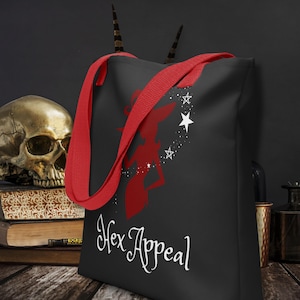 Puede incluir: Bolso tote negro con asas rojas. Presenta una silueta de bruja roja, estrellas y el texto "Hex Appeal" en escritura blanca. El bolso está hecho de un material duradero.