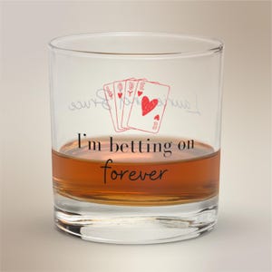 May include: A clear glass tumbler filled with amber liquid, featuring a poker-themed design. The glass has the text "I'm betting on forever" and a drawing of playing cards with hearts. The glass is suitable for whiskey or other spirits.