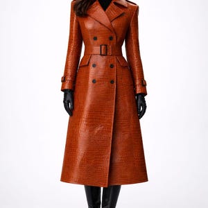 May include: A long, double-breasted, rust-colored trench coat with a textured, crocodile-like pattern. The coat features a belt, black buttons, and black gloves. The model is wearing black boots. The coat is a stylish outerwear garment.