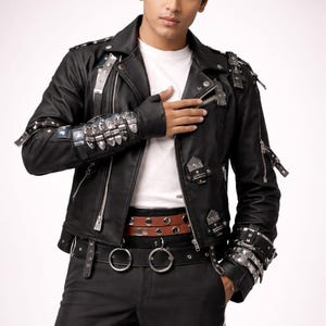 Puede incluir: Chaqueta de cuero negro con detalles metálicos plateados, cremalleras y hebillas. La chaqueta se combina con pantalones negros, un cinturón marrón y una camiseta blanca. El estilo es atrevido y moderno.