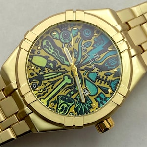May include: A gold-tone wristwatch with a metal link bracelet and a round case. The dial features a vibrant, multi-colored graphic print with various shapes and symbols, including a date window at the three o'clock position and the text Maurice Lacroix.