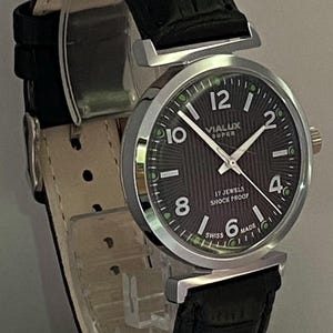 May include: A vintage VIALUX SUPER wristwatch with a black dial and silver case. The watch has white numerals, hands, and a black leather strap. The dial reads "17 JEWELS SHOCK PROOF SWISS MADE".