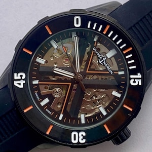May include: A Ulysse Nardin skeleton watch with a black rubber strap and a black bezel featuring white numerals at 0, 15, 30, and 45. The dial displays exposed mechanical movement components, orange accents, and white luminous markers on a dark background.