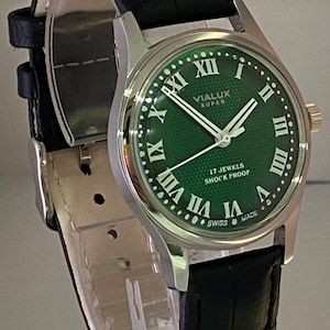 May include: A silver wristwatch with a green dial and a black leather strap. The watch face displays Roman numerals and the text "VIALUX SUPER", "17 JEWELS SHOCK PROOF", and "SWISS MADE".