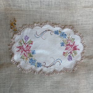 May include: An oval white fabric doily featuring a cross-stitch floral pattern in shades of pink, blue, purple, and green. The edges are finished with a scalloped beige crochet lace border. A small sprig of dried yellow flowers sits to the left.