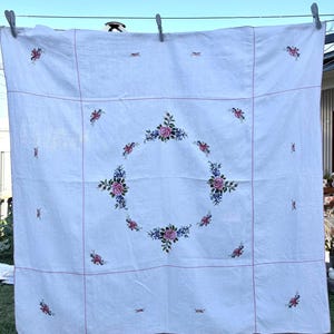 May include: A square white tablecloth featuring a central circular floral embroidery pattern of pink roses and blue flowers. The fabric is divided by thin red stitched lines, with smaller floral motifs placed in the corners and along the borders.