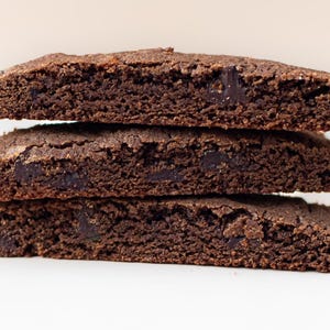 Puede incluir: Tres galletas rectangulares de color marr&oacute;n oscuro apiladas. Las galletas tienen una superficie rugosa y texturizada y parecen tener trozos de chocolate negro. El fondo es blanco.