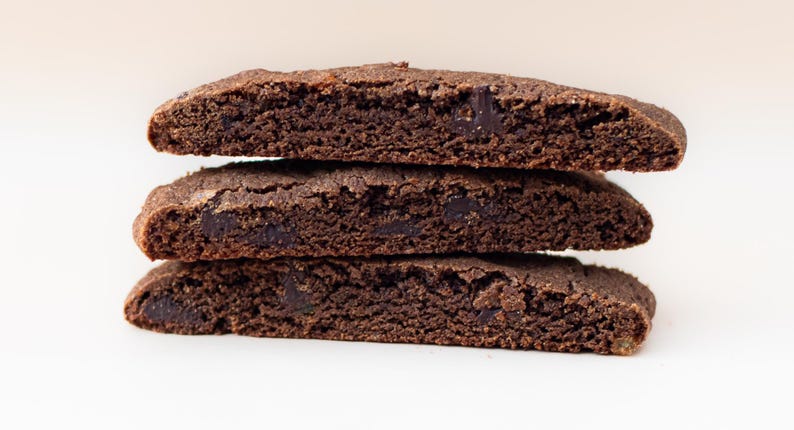 Puede incluir: Tres galletas rectangulares de color marr&oacute;n oscuro apiladas. Las galletas tienen una superficie rugosa y texturizada y parecen tener trozos de chocolate negro. El fondo es blanco.