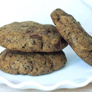 May include: Three round, golden-brown chocolate chip cookies with visible dark chocolate chunks and a textured surface. Two cookies are stacked horizontally, while the third cookie leans against them on a white, scalloped-edge plate.