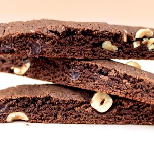 Puede incluir: Una pila de tres galletas biscotti de chocolate con avellanas visibles. Las galletas son de color marr&oacute;n oscuro con una superficie texturizada y chispas de chocolate visibles. Las avellanas son visibles en la parte superior y los lados.