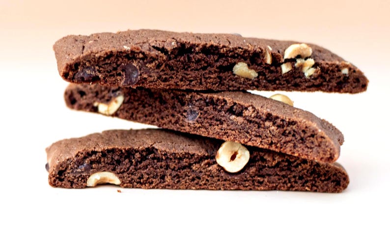 Puede incluir: Una pila de tres galletas biscotti de chocolate con avellanas visibles. Las galletas son de color marr&oacute;n oscuro con una superficie texturizada y chispas de chocolate visibles. Las avellanas son visibles en la parte superior y los lados.