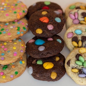 May include: An assortment of freshly baked cookies. Some are light-colored with colorful sprinkles, others are chocolate with candy-coated chocolates. A few cookies have chocolate chips and candy-coated chocolates. The cookies are arranged on a white surface.