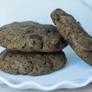 May include: Three chocolate chip cookies on a white, scalloped-edge plate. The cookies are a dark brown color with visible chocolate chips. One cookie leans against the other two. The plate is on a light-colored surface.