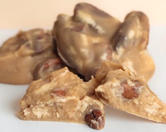 May include: A collection of light brown pecan pralines with a smooth, glossy surface and visible nut pieces embedded throughout. The confection has a dense, creamy texture with a broken piece in the foreground revealing a grainy interior.