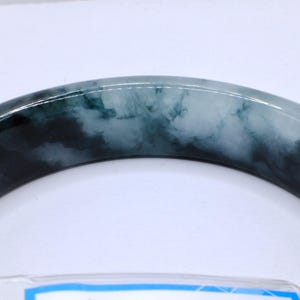 May include: A jade bangle bracelet with a marbled appearance. The bracelet features a mix of dark green and light green hues, creating a cloudy, natural pattern. The polished surface reflects light, highlighting the variations in color and texture.