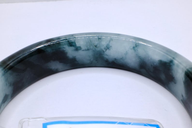 May include: A jade bangle bracelet with a marbled appearance. The bracelet features a mix of dark green and light green hues, creating a cloudy, natural pattern. The polished surface reflects light, highlighting the variations in color and texture.