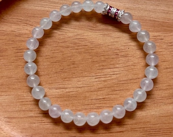 May include: A beaded bracelet composed of smooth, translucent pale pink round beads. The bracelet features a single cylindrical accent bead made of silver-toned metal, inlaid with three small, vibrant red stones and decorative filigree edges.