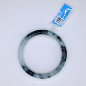 May include: A jade bangle bracelet in shades of green and black. The circular bracelet is displayed on a white background. A blue tag is attached to the bracelet.