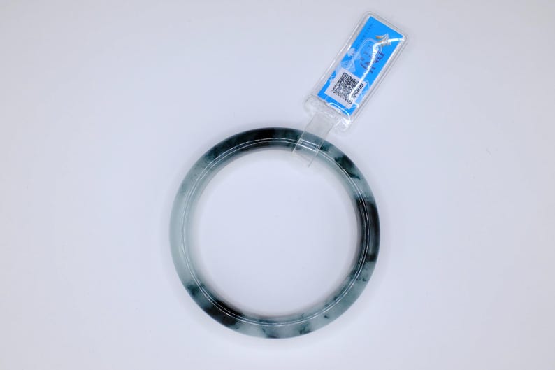 May include: A jade bangle bracelet in shades of green and black. The circular bracelet is displayed on a white background. A blue tag is attached to the bracelet.