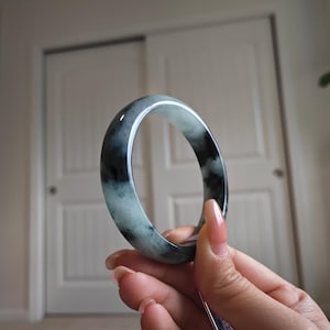 May include: A jade bangle bracelet with a mix of dark green and light green hues. The polished surface reflects light, showcasing the natural variations in the stone. The bracelet is held up against a neutral background.