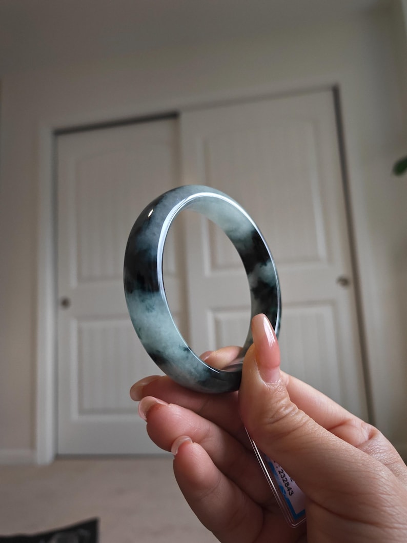 May include: A jade bangle bracelet with a mix of dark green and light green hues. The polished surface reflects light, showcasing the natural variations in the stone. The bracelet is held up against a neutral background.