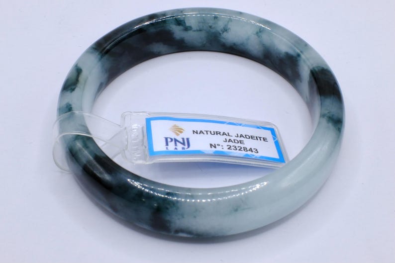 May include: A jade bangle bracelet with a marbled appearance, featuring shades of green and white. The bracelet has a smooth, polished surface. A tag with the text "NATURAL JADEITE JADE" and the number "232843" is attached.