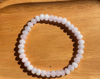 May include: A circular bracelet composed of numerous small, smooth, pale pink beads. The beads are uniform in size and shape, strung together to form a continuous loop. The light pink color of the beads is consistent throughout the entire piece.