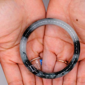 May include: A jade bangle bracelet with a gradient of black and white. The bracelet is held in open hands, with two rings visible on the fingers. The jade is polished and smooth, with a natural stone appearance.