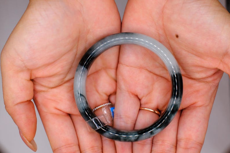 May include: A jade bangle bracelet with a gradient of black and white. The bracelet is held in open hands, with two rings visible on the fingers. The jade is polished and smooth, with a natural stone appearance.