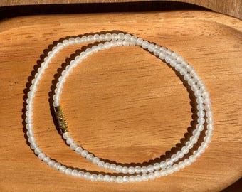 May include: A delicate necklace composed of small, translucent white beads arranged in a circular pattern. The strand features a cylindrical gold-toned metal clasp with a textured, knurled surface, providing a secure closure for the beaded jewelry piece.