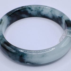 May include: A jade bangle bracelet with a marbled appearance. The bracelet features shades of dark green and white, creating a natural, flowing pattern. The smooth, polished surface reflects light, highlighting the color variations. A tag is attached.