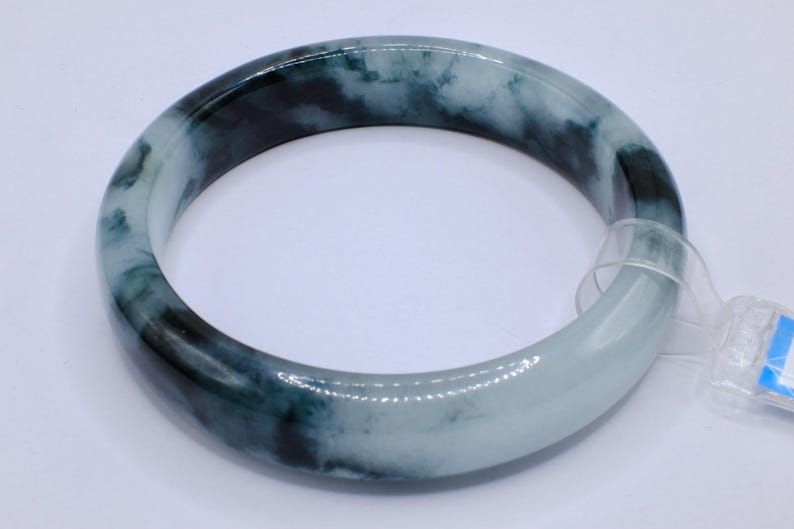 May include: A jade bangle bracelet with a marbled appearance. The bracelet features shades of dark green and white, creating a natural, flowing pattern. The smooth, polished surface reflects light, highlighting the color variations. A tag is attached.