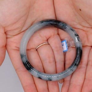 May include: A jade bangle bracelet with a mix of gray and black tones. The bracelet is round and smooth, with a polished finish. The jade is held in open hands, and a small ring is visible on one finger. A small tag is attached to the bracelet.