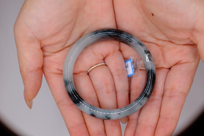 May include: A jade bangle bracelet with a mix of gray and black tones. The bracelet is round and smooth, with a polished finish. The jade is held in open hands, and a small ring is visible on one finger. A small tag is attached to the bracelet.