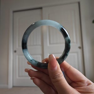 May include: A jade bangle bracelet with a smooth, polished surface. The bracelet features a blend of light blue and dark green hues, creating a marbled effect. The circular design is held up in front of a white background.