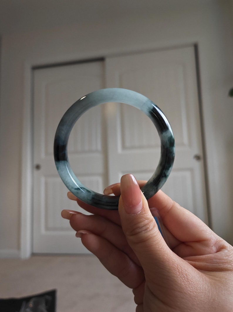 May include: A jade bangle bracelet with a smooth, polished surface. The bracelet features a blend of light blue and dark green hues, creating a marbled effect. The circular design is held up in front of a white background.