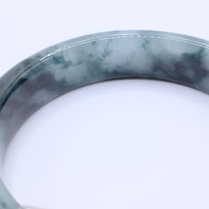 May include: A jade bangle bracelet with a marbled appearance. The bracelet features shades of green and gray, with a polished, smooth surface. The bangle is a circular shape, and the image is set against a white background.