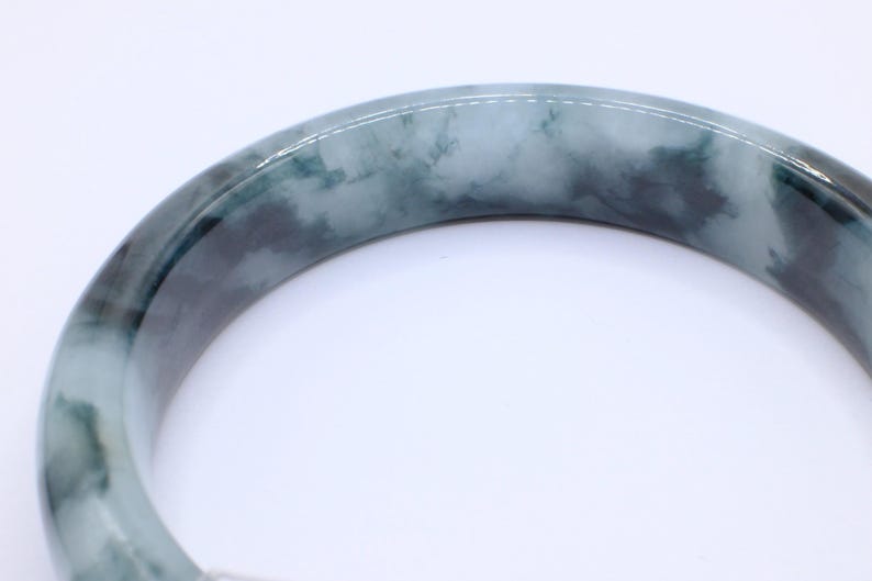 May include: A jade bangle bracelet with a marbled appearance. The bracelet features shades of green and gray, with a polished, smooth surface. The bangle is a circular shape, and the image is set against a white background.