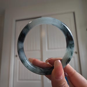 May include: A jade bangle bracelet with a marbled appearance. The bracelet features shades of green and black, with a polished, glossy finish. The circular bracelet is held up by a hand.