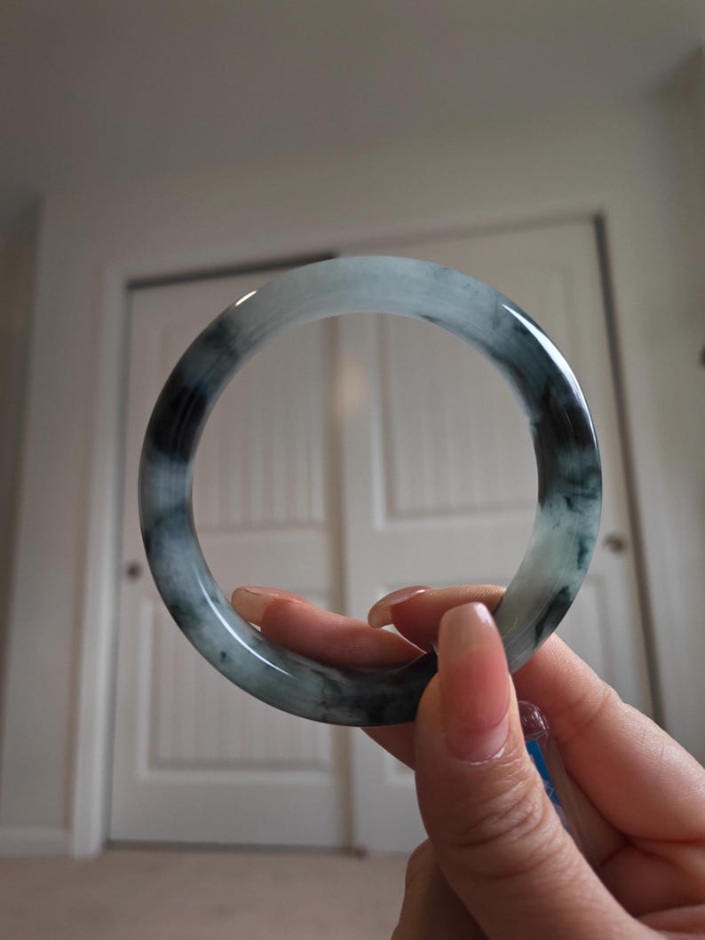 May include: A jade bangle bracelet with a marbled appearance. The bracelet features shades of green and black, with a polished, glossy finish. The circular bracelet is held up by a hand.