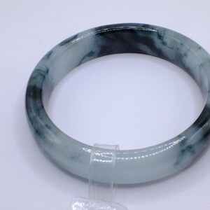 May include: A jade bangle bracelet with a marbled appearance. The bracelet features shades of light green and dark green, creating a natural, organic pattern. The smooth, polished surface reflects light, highlighting the variations in color and texture. This piece is a classic jewelry item.