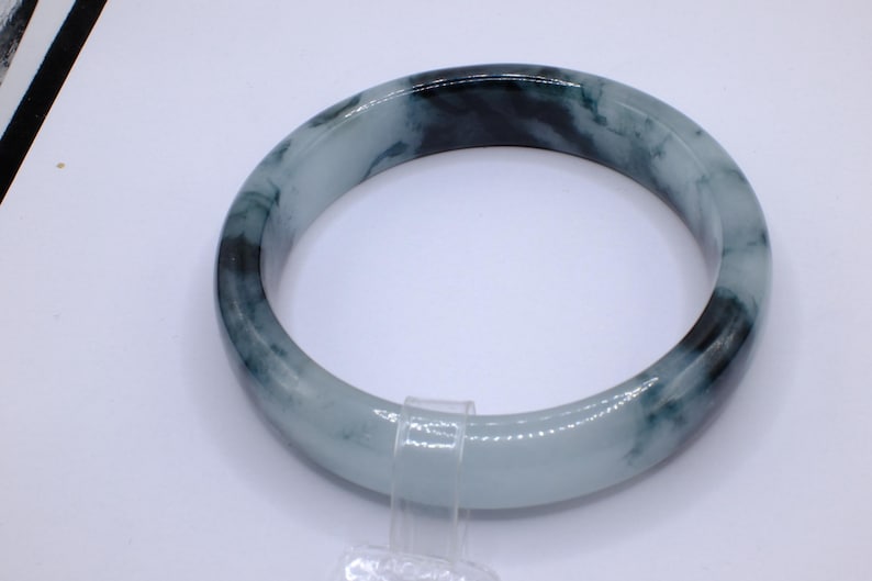 May include: A jade bangle bracelet with a marbled appearance. The bracelet features shades of light green and dark green, creating a natural, organic pattern. The smooth, polished surface reflects light, highlighting the variations in color and texture. This piece is a classic jewelry item.
