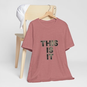 Puede incluir: Una camiseta rosa polvoriento con las palabras "THIS IS IT" en una fuente texturizada negra y dorada. La camiseta está colgada en una percha de madera. Una gorra de béisbol blanca descansa sobre el respaldo de una silla de madera.