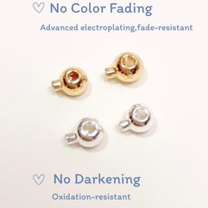 Puede incluir: Dos cuentas esféricas doradas y dos plateadas con pequeños bucles. El texto dice: No Color Fading, Advanced electroplating, fade-resistant, No Darkening, Oxidation-resistant. Estas cuentas metálicas presentan un acabado liso y reflectante.